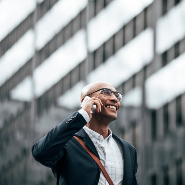 Smiling businessman talking over cell phone while commuting to office with a glass facade building in background. Man in formal clothes carrying office bag walking on street while talking on mobile phone.